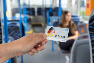 Hand hält ein D-Ticket im Bus, Fahrgast im Hintergrund.